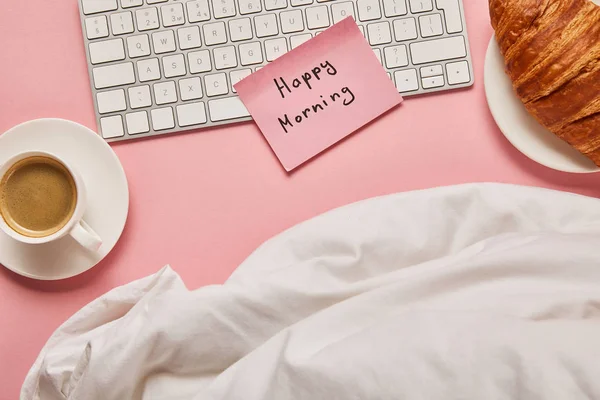 Top view of blanket, computer keyboard and pink sticky note with happy morning lettering near croissant and coffee on pink background — Stock Photo