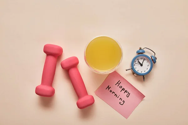 Vista superior de la nota adhesiva con letras de la mañana feliz, mancuernas de color rosa, jugo de naranja y pequeño reloj despertador sobre fondo beige - foto de stock