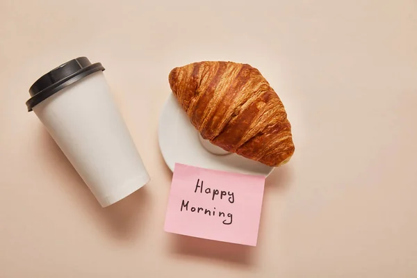 Vista superior del café para llevar, croissant y nota adhesiva con letras de la mañana feliz sobre fondo beige - foto de stock