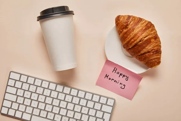 Vue du haut du café à emporter, croissant, clavier d'ordinateur et note collante avec lettrage du matin heureux sur fond beige — Photo de stock