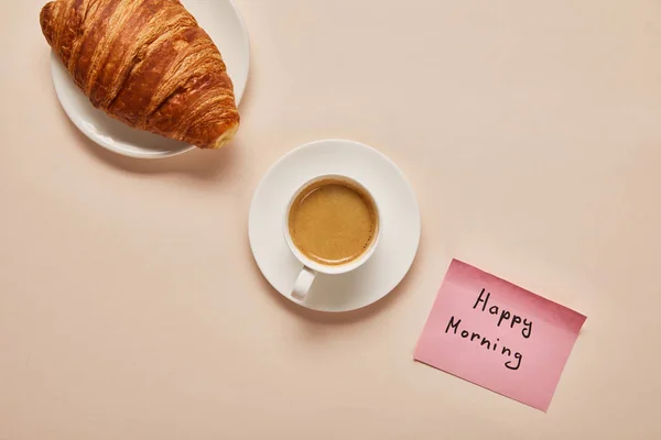 Plat étendu avec café, croissant et note collante avec lettrage matin heureux sur fond beige — Photo de stock