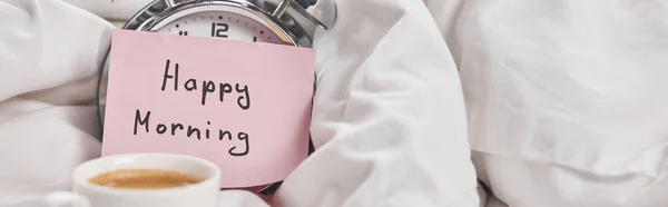 Coffee in white cup on saucer near silver alarm clock with happy morning lettering on sticky note in bed, panoramic shot — Stock Photo