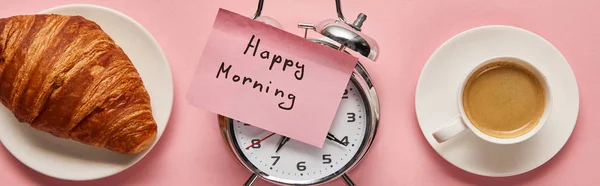 Vista superior do despertador com letras da manhã feliz na nota pegajosa perto de café e croissant no fundo rosa, tiro panorâmico — Fotografia de Stock