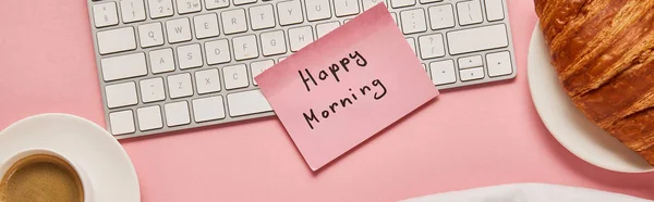Top view of computer keyboard and pink sticky note with happy morning lettering near croissant and coffee on pink background, panoramic shot — Stock Photo