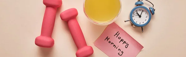 Top view of sticky note with happy morning lettering, pink dumbbells, orange juice and small alarm clock on beige background, panoramic shot — Stock Photo