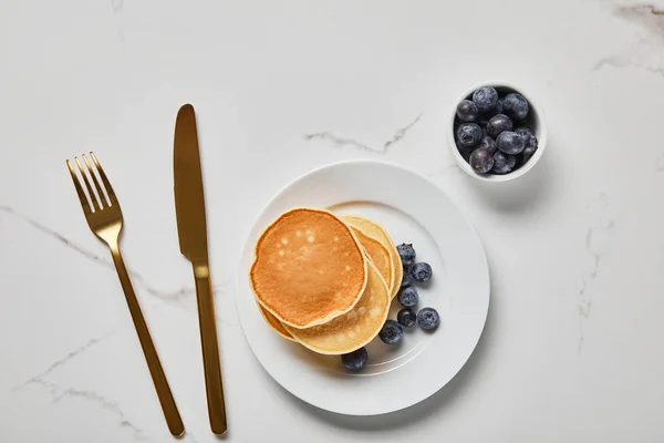 Top view of pancakes on plate near golden cutlery and bowl with blueberries — Stock Photo