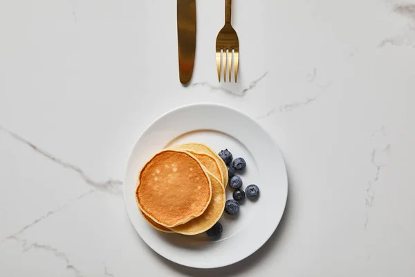 Top view of pancakes with blueberries on plate near golden fork and knife — Stock Photo