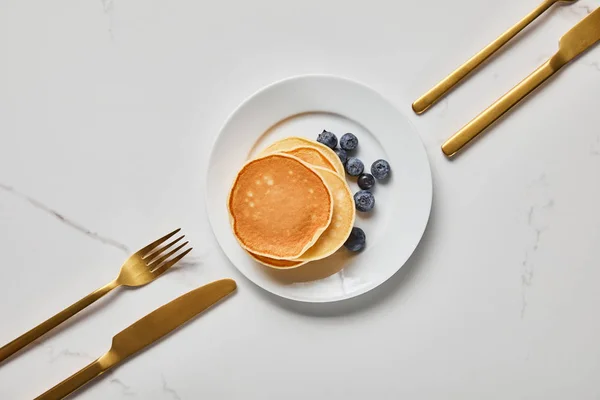 Top view of plate with pancakes and blueberries near forks and knives — Stock Photo