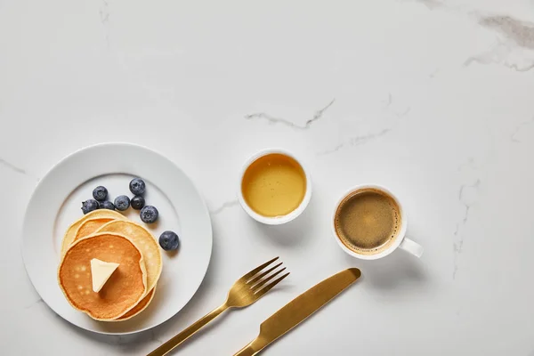 Vista dall'alto della ciotola con miele, gustose frittelle con mirtilli e tazza di caffè vicino a posate dorate — Stock Photo