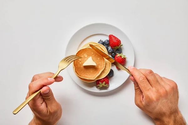 Abgeschnittene Ansicht eines Mannes, der Pfannkuchen auf Teller mit Blaubeeren und Erdbeeren schneidet — Stockfoto