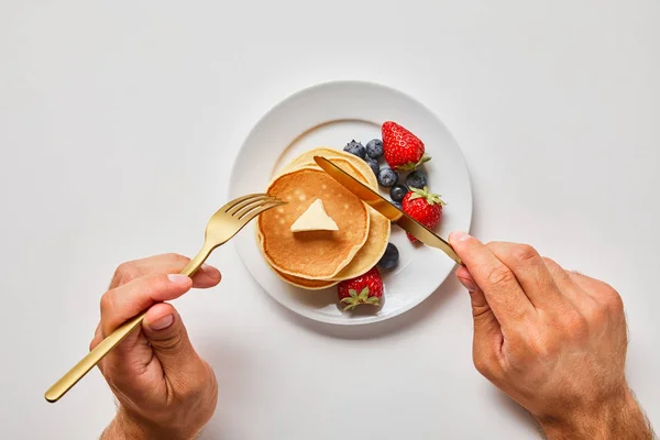 Cropped view of man cutting pancakes on plate with blueberries and strawberries — Stock Photo