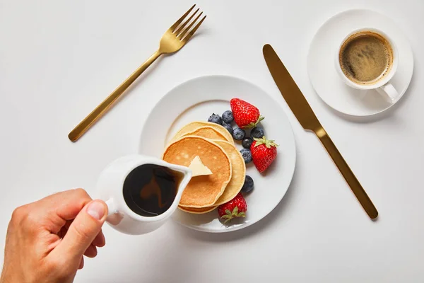 Vista cortada do homem derramando xarope em panquecas com bagas perto de talheres dourados e xícara de café — Fotografia de Stock