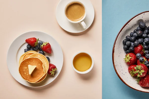 Top view of pancakes with berries on plate, bowl with honey, cup of coffee and plate with berries on pink and blue background — Stock Photo