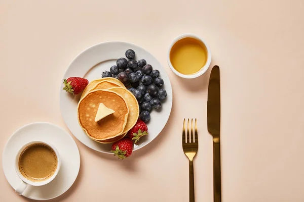 Vista superior del desayuno con taza de café, panqueques, fresas, arándanos y miel cerca de cubiertos de oro - foto de stock