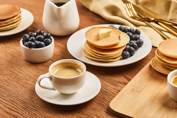 Gustosa colazione con tazza di caffè, pancake e mirtilli vicino al panno di lino con posate su superficie di legno — Foto stock