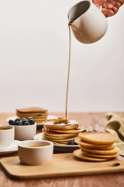 Abgeschnittene Ansicht eines Mannes, der leckere Pfannkuchen mit Sirup aus einer Kanne in der Nähe von Schüsseln mit Blaubeeren und Honig gießt, und eine Tasse Kaffee isoliert auf grau — Stockfoto