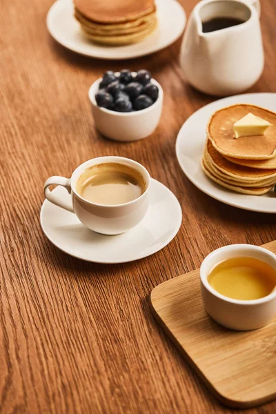 Selektiver Fokus der Tasse Kaffee auf Untertasse neben Teller mit Pfannkuchen und Schalen mit Honig und Blaubeeren auf Holzoberfläche — Stockfoto