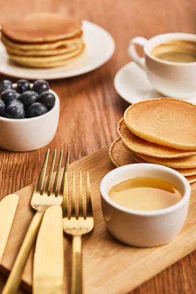 Selektiver Fokus der Schüssel mit Honig, Pfannkuchen und Blaubeeren in der Nähe einer Tasse Kaffee auf Holzoberfläche — Stockfoto