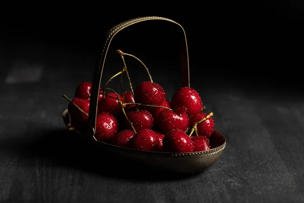 Rojo deliciosas cerezas húmedas en cesta de metal sobre mesa oscura de madera aislada en negro - foto de stock