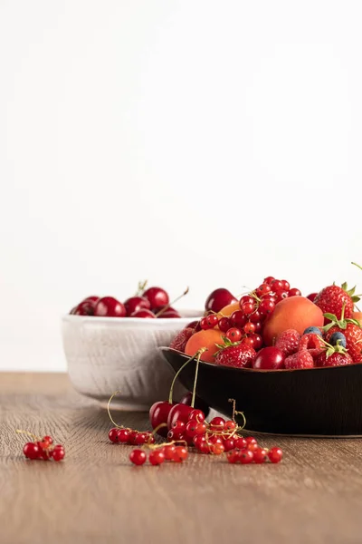 Enfoque selectivo de cerezas rojas en tazón blanco y bayas mixtas en plato en mesa de madera aislada en blanco - foto de stock