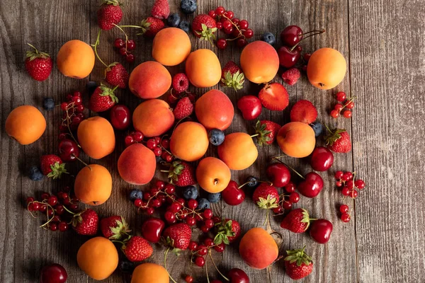 Blick von oben auf reife köstliche frische Beeren und Aprikosen auf Holztisch verstreut — Stockfoto