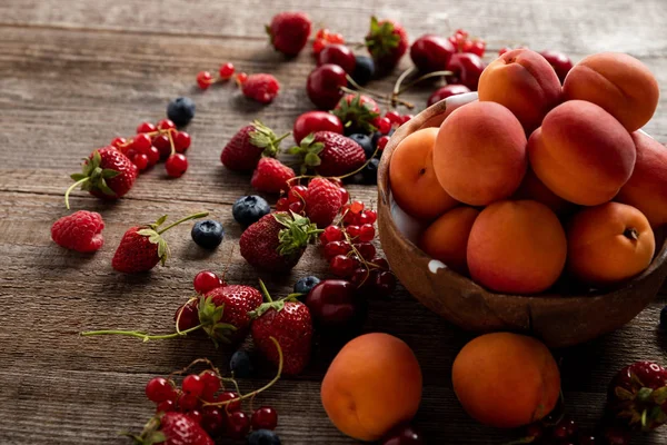 Спелые вкусные сезонные ягоды, разбросанные по чаше со свежими абрикосами на деревянном столе — стоковое фото