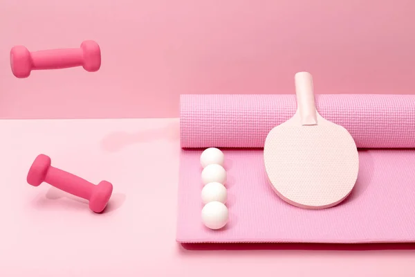 Pink dumbbells levitating in air near white ping-pong balls and racket on fitness mat on pink background — Stock Photo