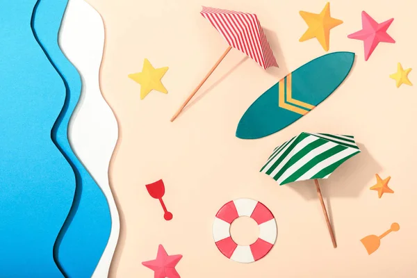 Верхний вид бумажного пляжа с летними зонтиками, спасательным буем и доской для серфинга на песке возле океана — стоковое фото