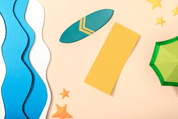 Верхний вид бумажного пляжа с желтым полотенцем, зеленым зонтиком и доской для серфинга рядом с морскими звездами на песке возле океана — стоковое фото