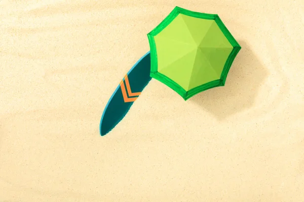 Вид сверху на бумажный пляж с зеленым зонтиком и доской для серфинга на текстурированном песке — стоковое фото