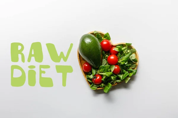 Top view of fresh green vegetables in heart shaped bowl on white background with raw diet lettering — Stock Photo