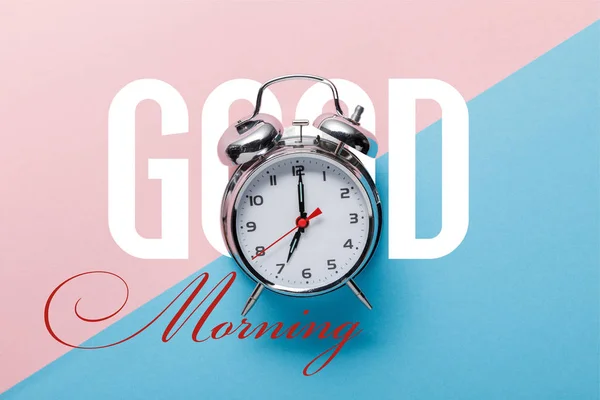 Vista superior del reloj despertador clásico plateado sobre fondo rosa y azul con letras de buena mañana - foto de stock