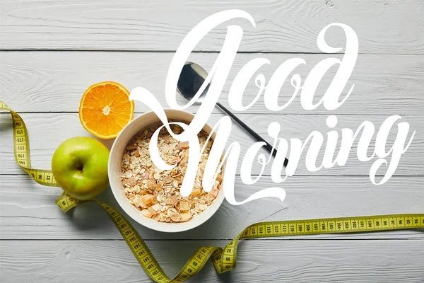 Vista superior de cinta métrica, cuchara y cereal de desayuno en tazón cerca de manzana y naranja sobre fondo blanco de madera con letras de buenos días — Stock Photo