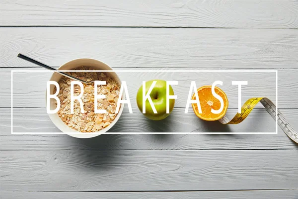 Plano con cereales para el desayuno en tazón, manzana, naranja y cinta métrica sobre fondo blanco de madera con letras de desayuno - foto de stock