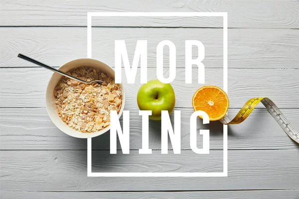 Plano con cereales para el desayuno en tazón, manzana, naranja y cinta métrica sobre fondo blanco de madera con letras de la mañana - foto de stock