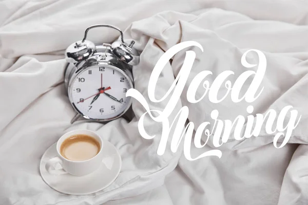 Coffee in white cup on saucer near silver alarm clock in bed with good morning illustration — Stock Photo