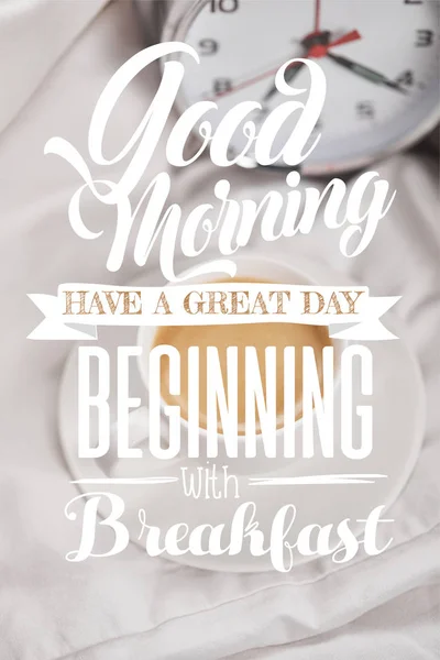 Top view of coffee in white cup on saucer near silver alarm clock on bedding with good morning, have a great day beginning with breakfast lettering — Stock Photo