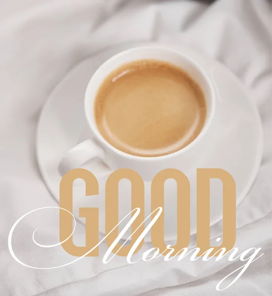 Top view of coffee in white cup on saucer near silver alarm clock on bedding with good morning illustration — Stock Photo