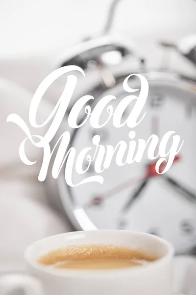 Selective focus of coffee in white cup with silver alarm clock in bed on background with good morning illustration — Stock Photo