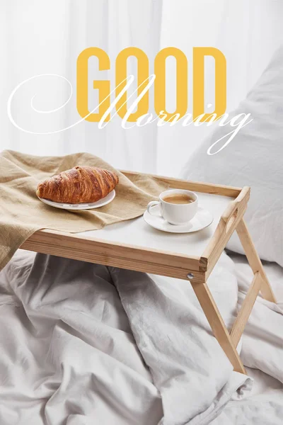 Кофе и круассан подается на деревянном подносе на белой кровати с подушкой с иллюстрацией доброго утра — стоковое фото