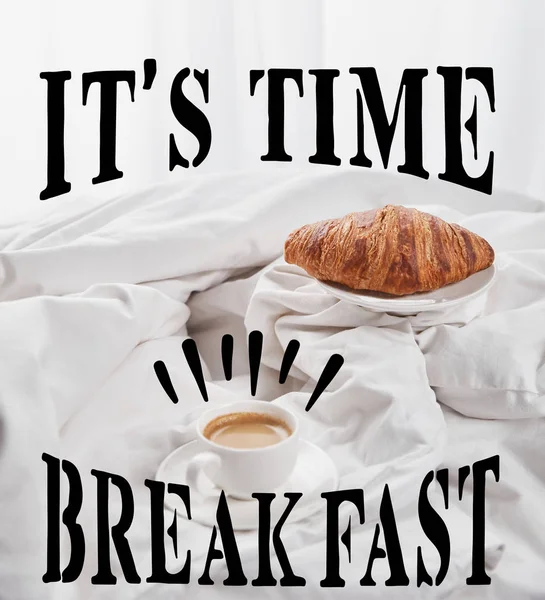 Frisches Croissant auf Teller neben Kaffee in weißer Tasse auf Untertasse im Bett mit seiner Zeit, Frühstücksschrift — Stockfoto
