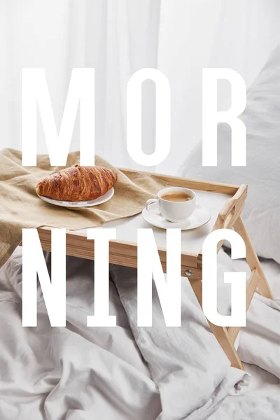 Кофе и круассан подается на деревянном подносе на белой кровати с подушкой с утренней иллюстрацией — стоковое фото
