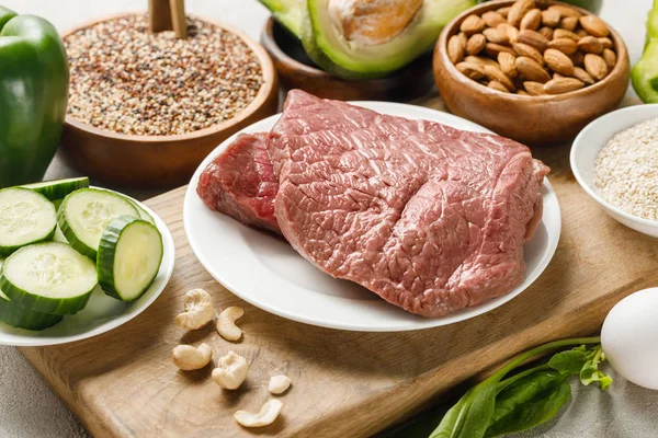 Carne cruda sobre tabla de cortar de madera cerca de nueces y verduras verdes, menú de dieta cetogénica — Stock Photo