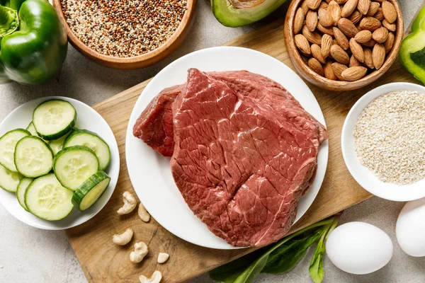 Vista superior de la carne cruda en la tabla de cortar de madera cerca de nueces, grañones, huevos y verduras verdes, menú de dieta cetogénica - foto de stock