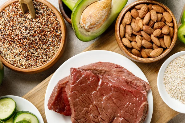 Вид на свежее сырое мясо на белой тарелке возле орехов, крупы и авокадо, кетогенное диетическое меню — стоковое фото
