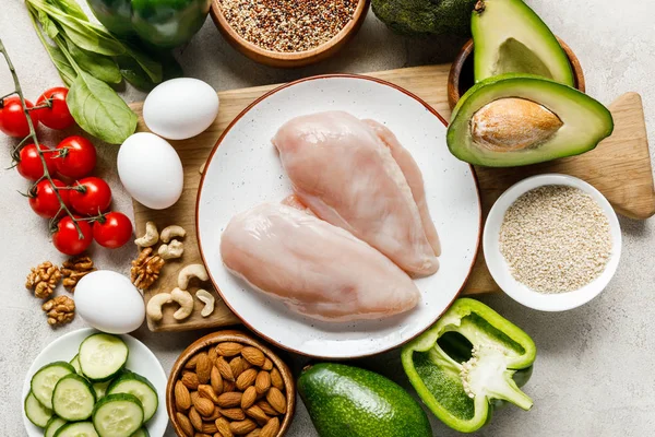 Vue du dessus des poitrines de poulet crues fraîches sur assiette blanche près des noix, des œufs et des légumes mûrs, menu diététique cétogène — Photo de stock