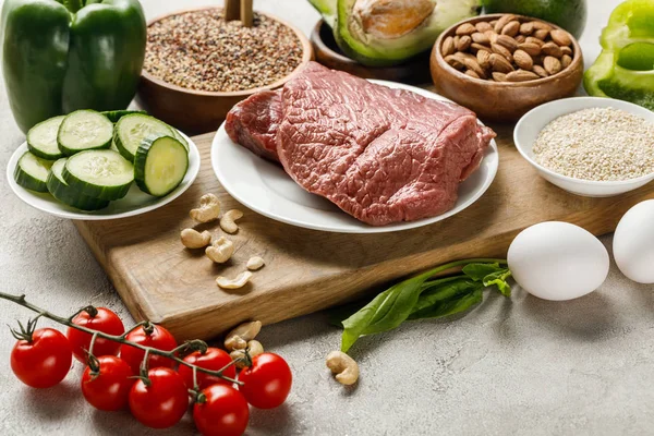 Carne fresca cruda sobre tabla de cortar de madera cerca de nueces, verduras y grañones, menú de dieta cetogénica - foto de stock