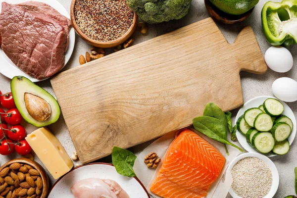 Ansicht von rohem Lachs, Hühnerbrust und Fleisch in der Nähe von Nüssen, Milchprodukten und Gemüse rund um ein hölzernes Schneidebrett mit Kopierraum, ketogene Speisekarte — Stockfoto