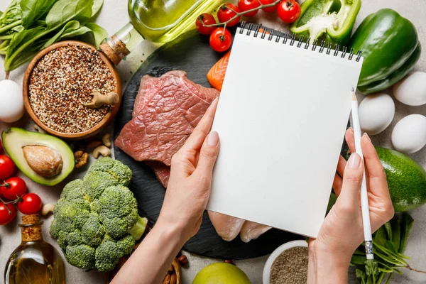 Vista cortada da mulher segurando notebook vazio acima de alimentos para menu de dieta cetogênica — Fotografia de Stock