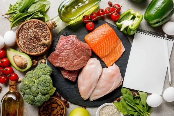 Вид зверху порожній блокнот біля сирого м'яса і риби серед свіжих овочів, кетогенне меню дієти — стокове фото